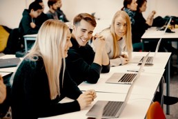 Foto av to kvinnelige og en mannlig student i et klasserom foran en pult med hver sin laptop.