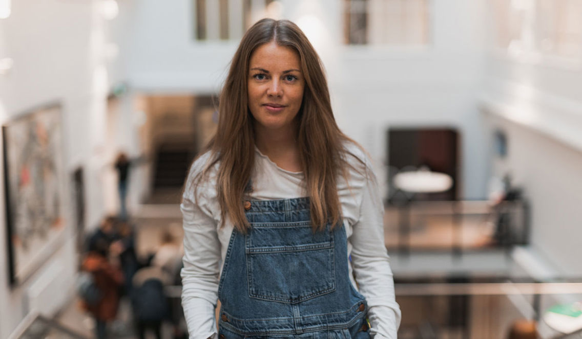 Rikke Gregersen poserer på Høyskolen Kristiania Fjerdingen. Hun har langt hår og har på seg snekkerbukser. 