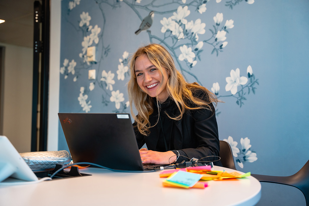 Bilde av Malin Voldberg som sitter ved en en kontorpult med en bærbar PC, og smiler til kamera.