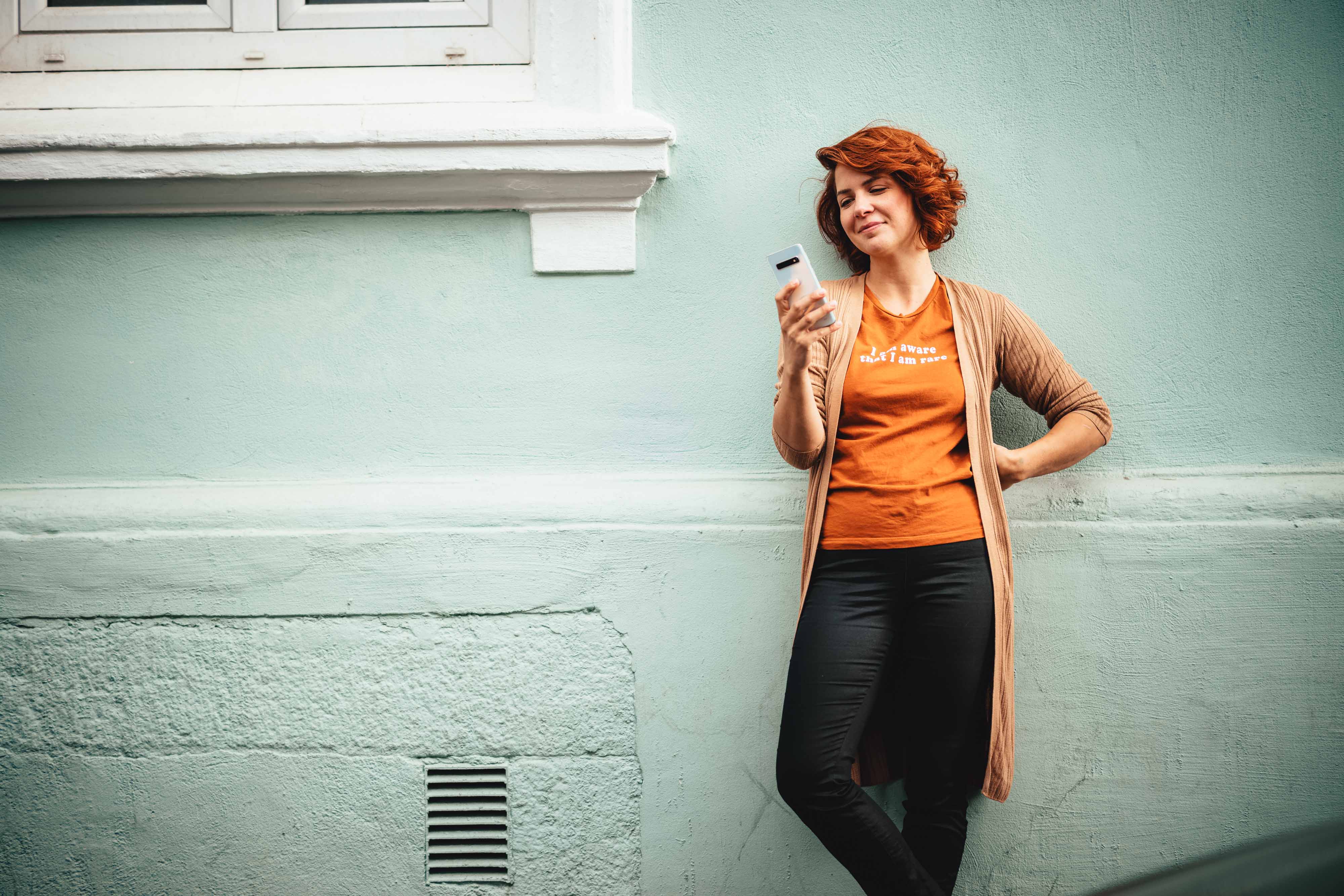 Foto av ung kvinne i oransje overdel som lener seg mot en husvegg med mobiltelefon i hånden.