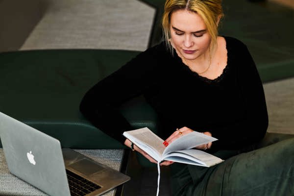 Ung blond kvinne i mørk genser leser bok ved siden av en laptop