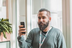 En blid mann med skjegg ser inn i kamera med en telefon i hånden. 