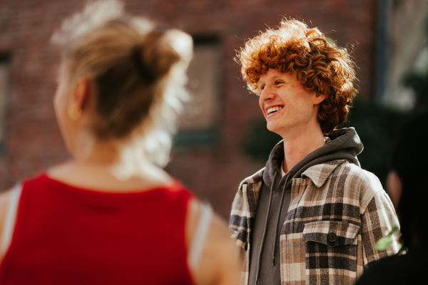 En ung person med krøllete rødt hår og et plaidjakke smiler mens de ser til venstre. En person med ryggen til kameraet, som bærer en rød topp ser samme retning.