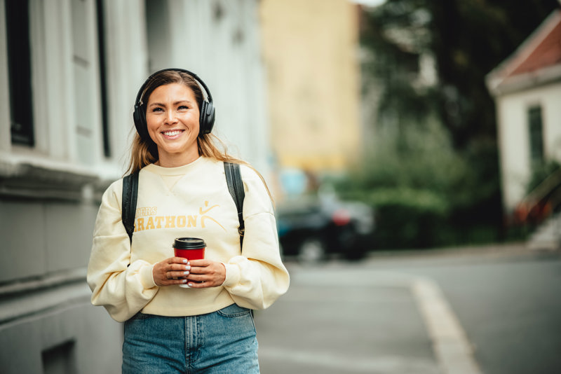 Foto av ung kvinne med hodetelefoner og kaffekopp som vandrer langs en gate.