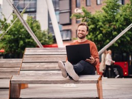 Foto av mann som sitter på en benk med beina høyt, og har en laptop i fanget.