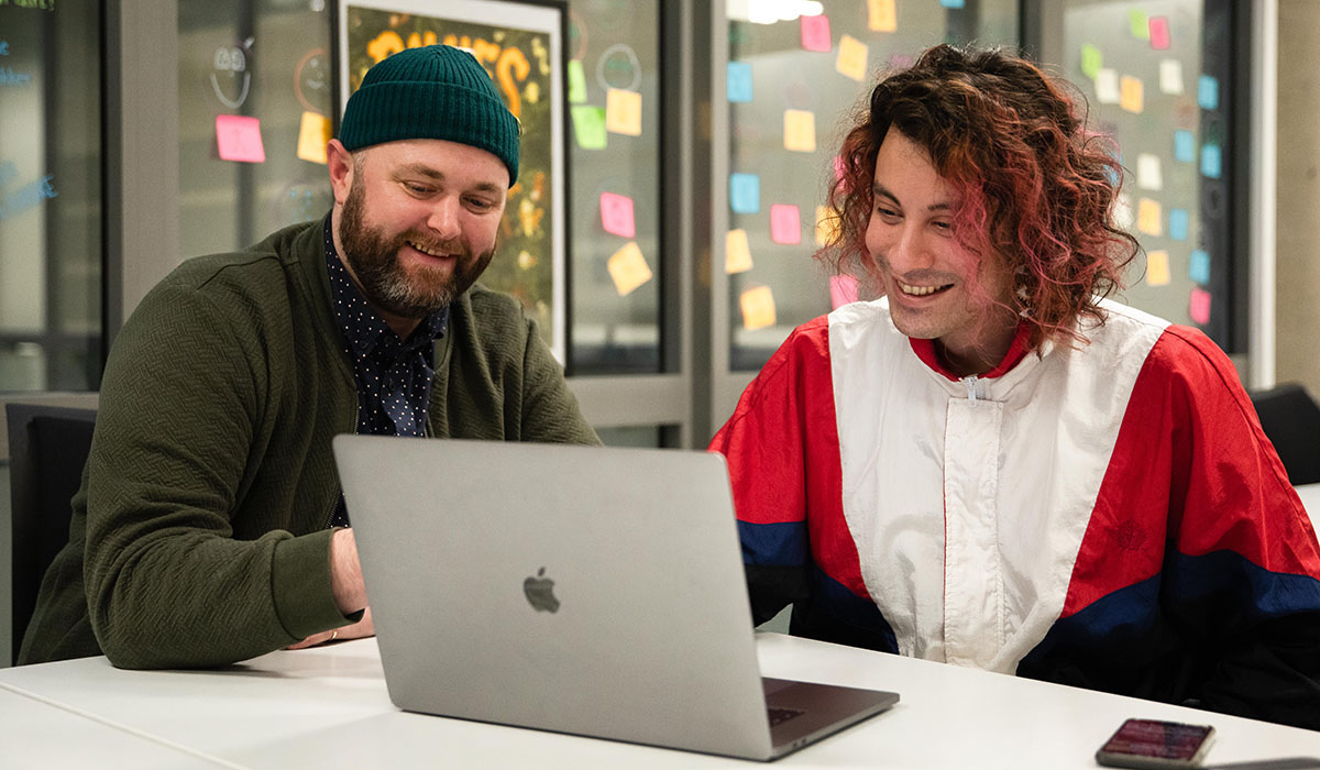 Foto av to menn som sitter foran en MacBook og smiler. 