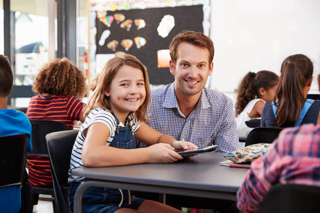 En smilende voksen og et barn ser på en nettbrett sammen i et klasserom. Bak dem er en tavle med fargerike kunstverk.