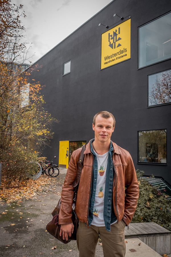 Ung mannlig skuespillerstudent foran Westerdals Høyskolen Kristiania på Vulkan. Det er høst, bladene på trærne bak ham er gule og oransje.