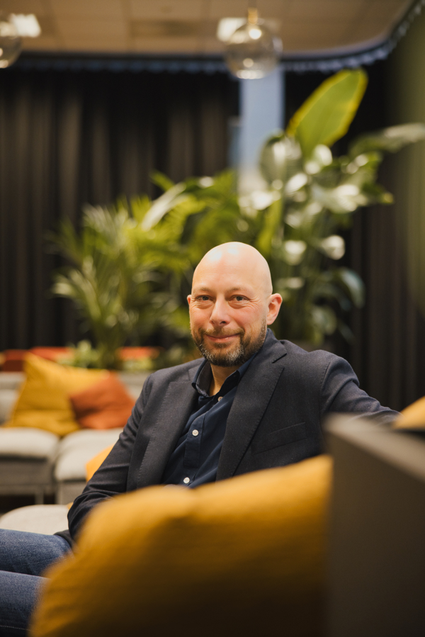 Portrettbilde av Tor-Morten Grønli som sitter i en grå sofa med brune puter og grønne planter i bakgrunnen